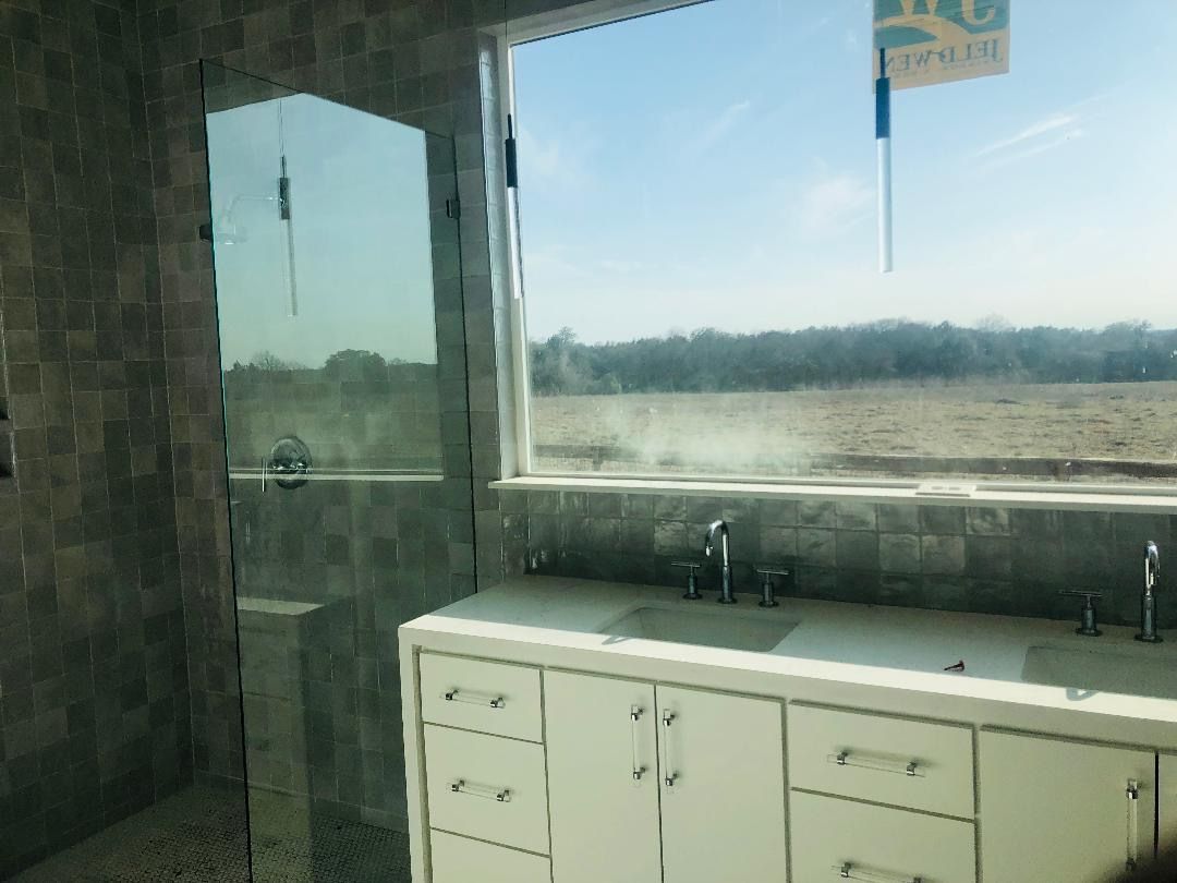 A bathroom vanity with white cabinets and faucets sits in front of a window looking out over a grassy field.