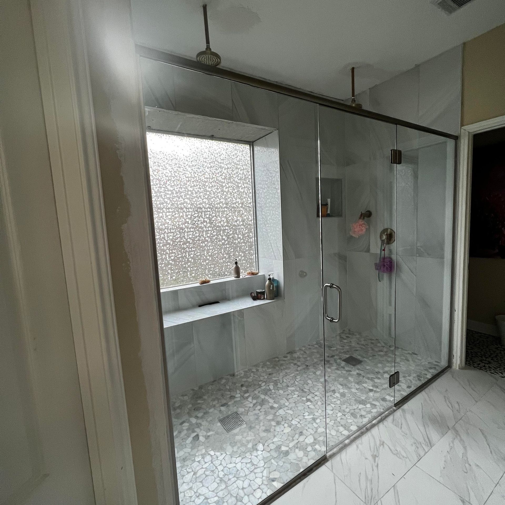 A modern walk-in shower with white marble tiles, a glass door, a rain shower head, and a window with a patterned film.