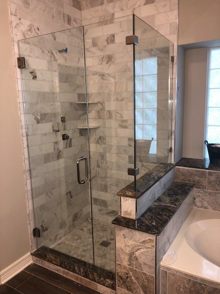 A modern shower with glass door, marble-tiled walls, and a granite-topped ledge next to a bathtub.