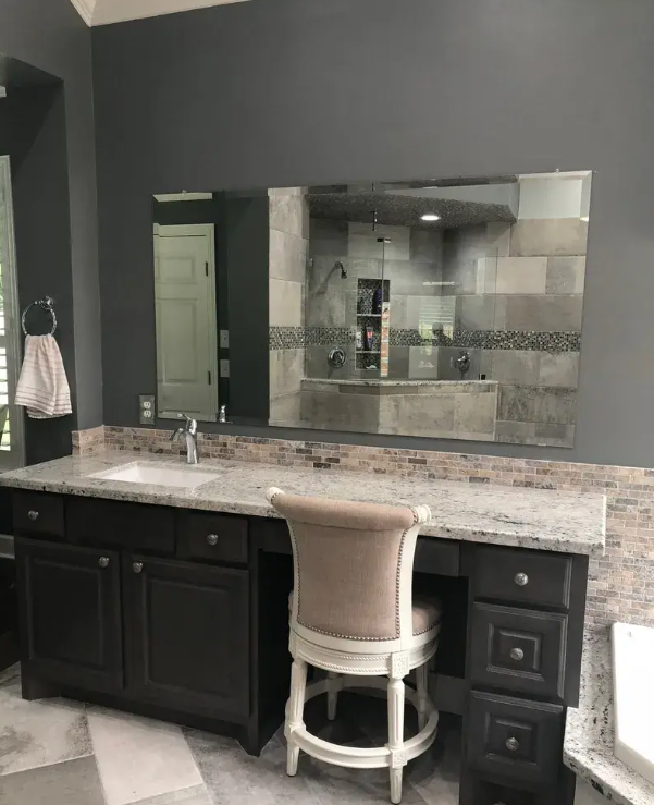 A dark-painted bathroom vanity with a speckled granite countertop, a single sink, a mirror, and a cushioned vanity chair.