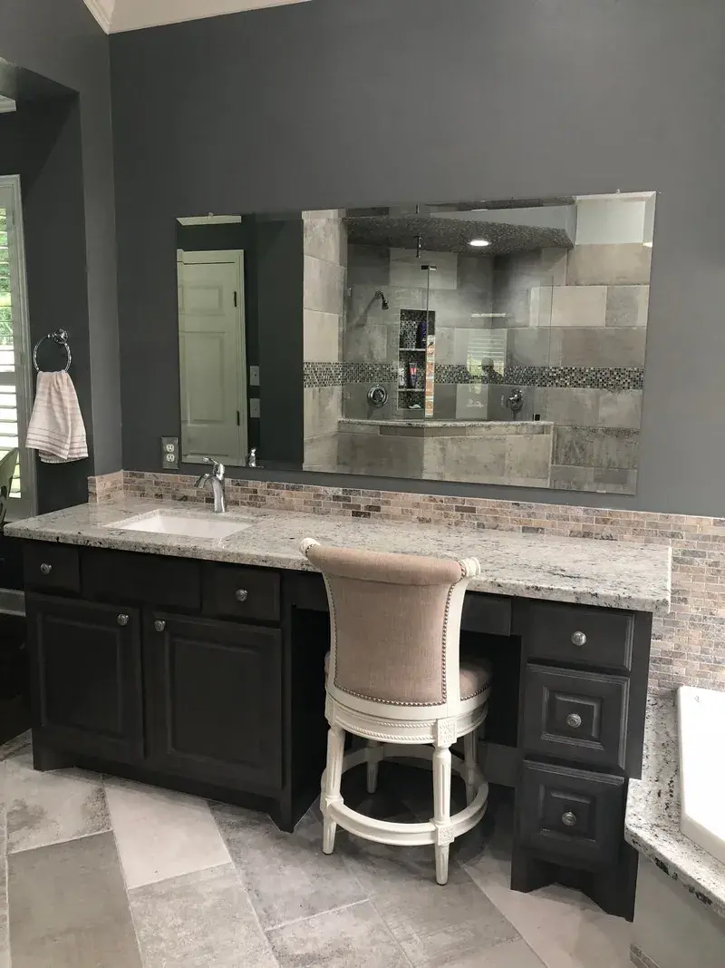 A dark gray bathroom vanity with granite countertop, large mirror, and a beige stool, set against a stone-tiled wall.