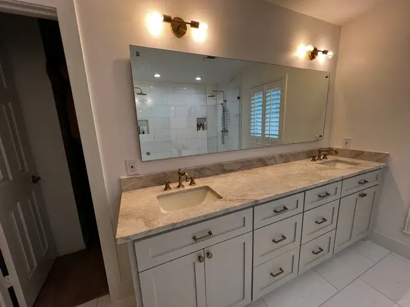 A bathroom vanity with a long stone countertop, double undermount sinks, white cabinets, a large mirror, and wall lights.