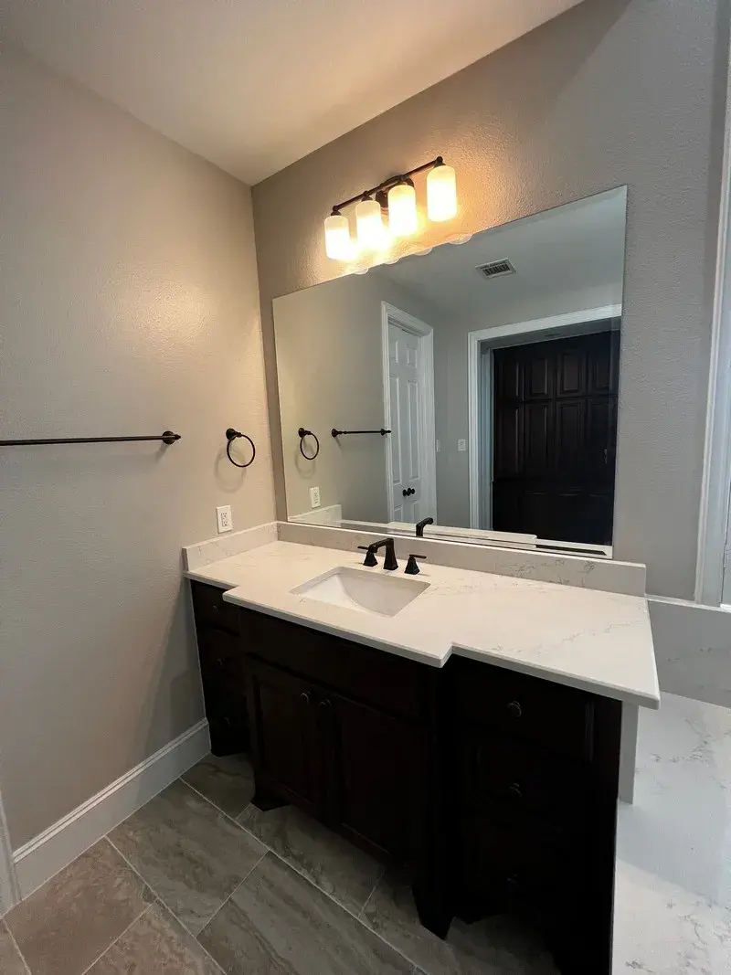 A modern bathroom vanity with dark wood cabinets, a white countertop, an oval mirror, and a three-light fixture.