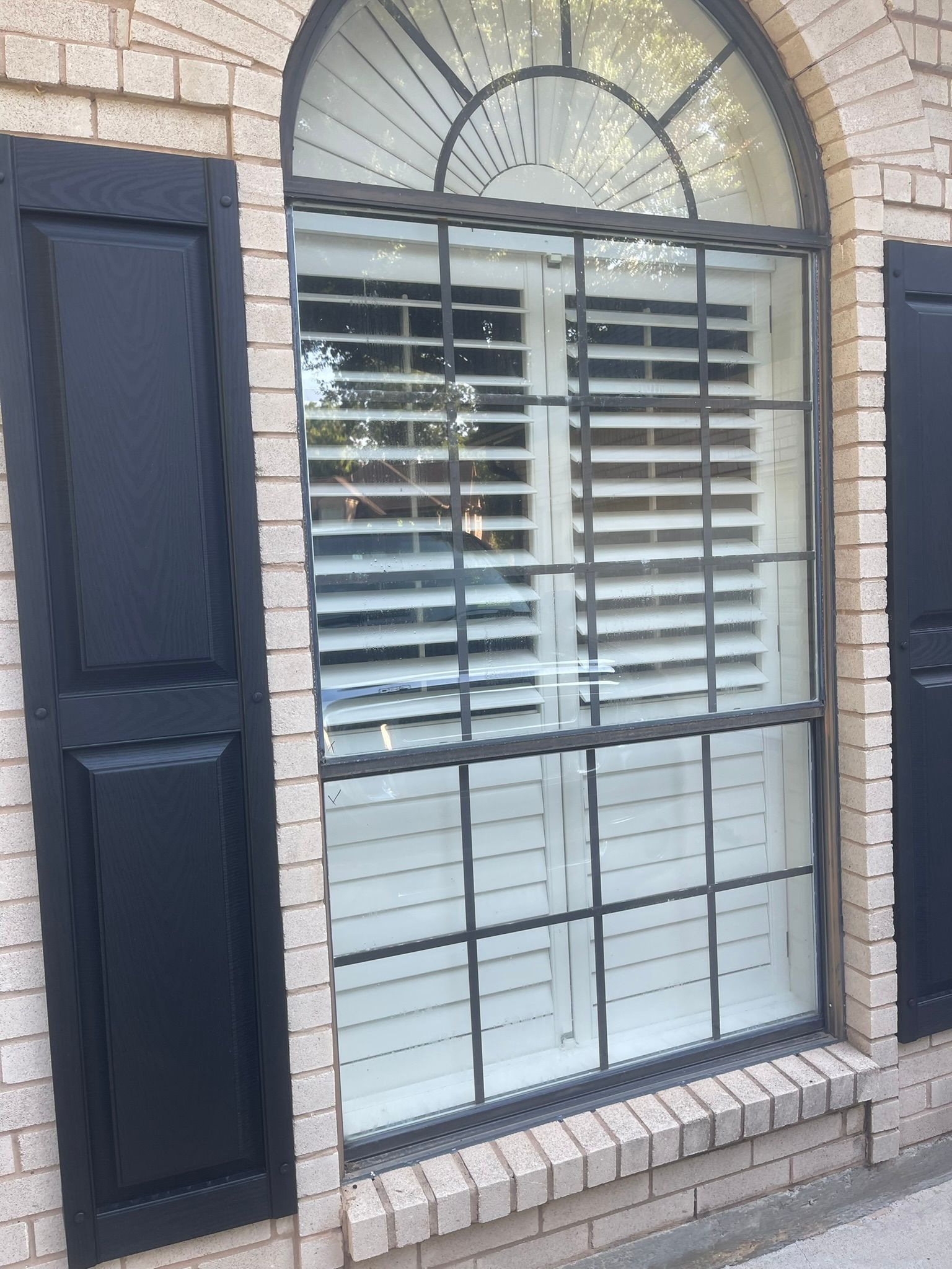 An arched window with dark shutters on a light brick wall, featuring white interior blinds and a metal grid overlay.