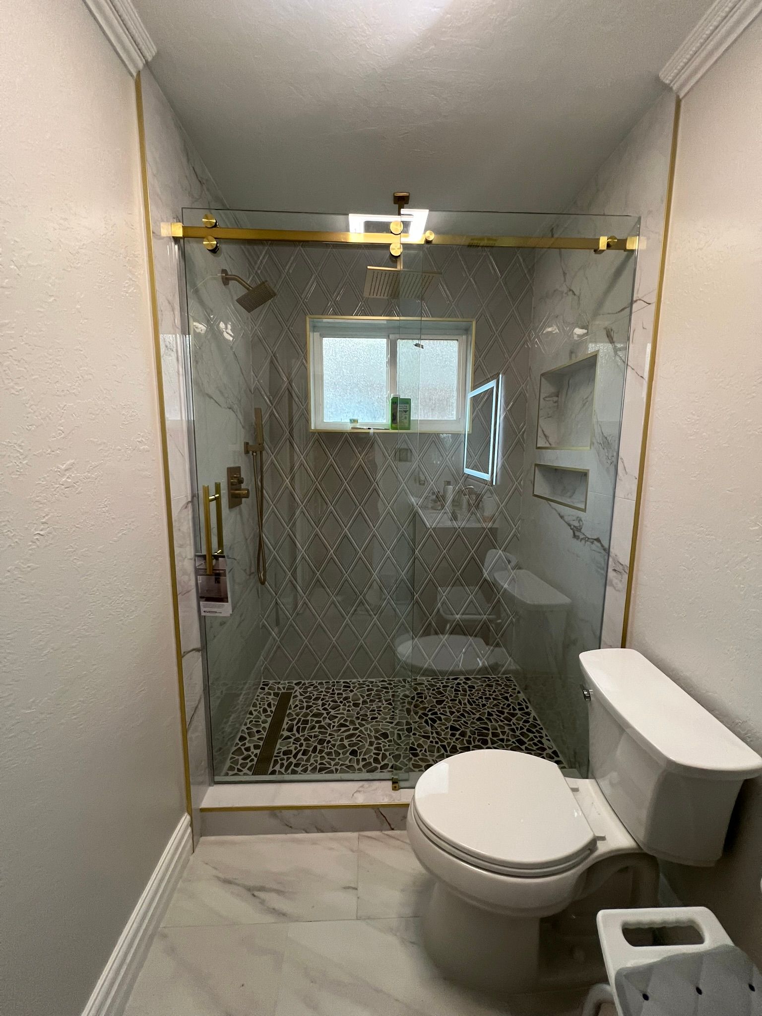 A bathroom featuring a white toilet, marble-patterned flooring, and a walk-in shower with gold trim and tiled walls.