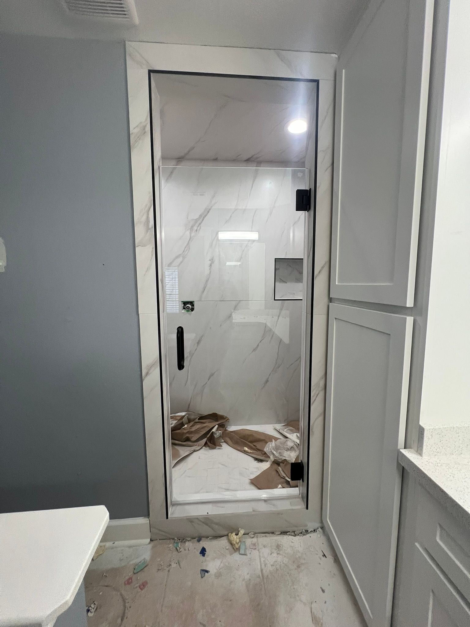 A glass shower door with a black frame and handle set against white marble-patterned tile in a bathroom under renovation.