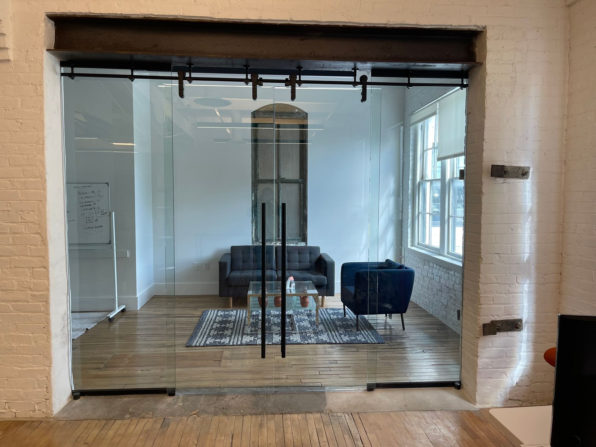 A glass wall with sliding doors leads into a small office containing a sofa, an armchair, a coffee table, and a rug.