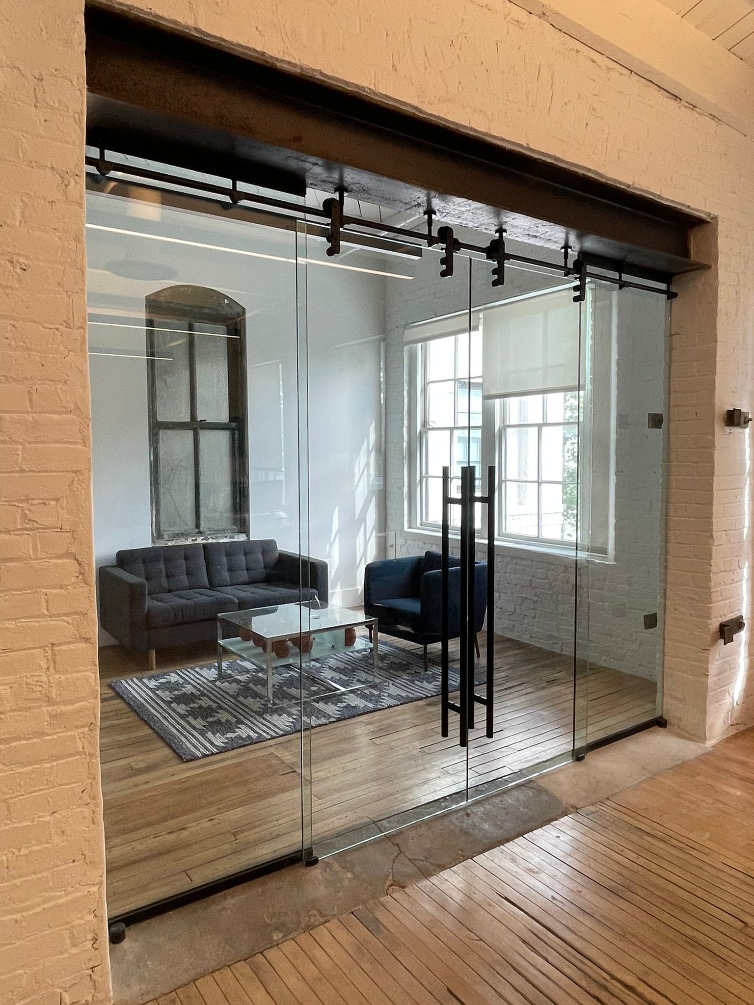 Glass sliding doors with dark metal hardware lead into a furnished office space with exposed brick walls and hardwood floors.