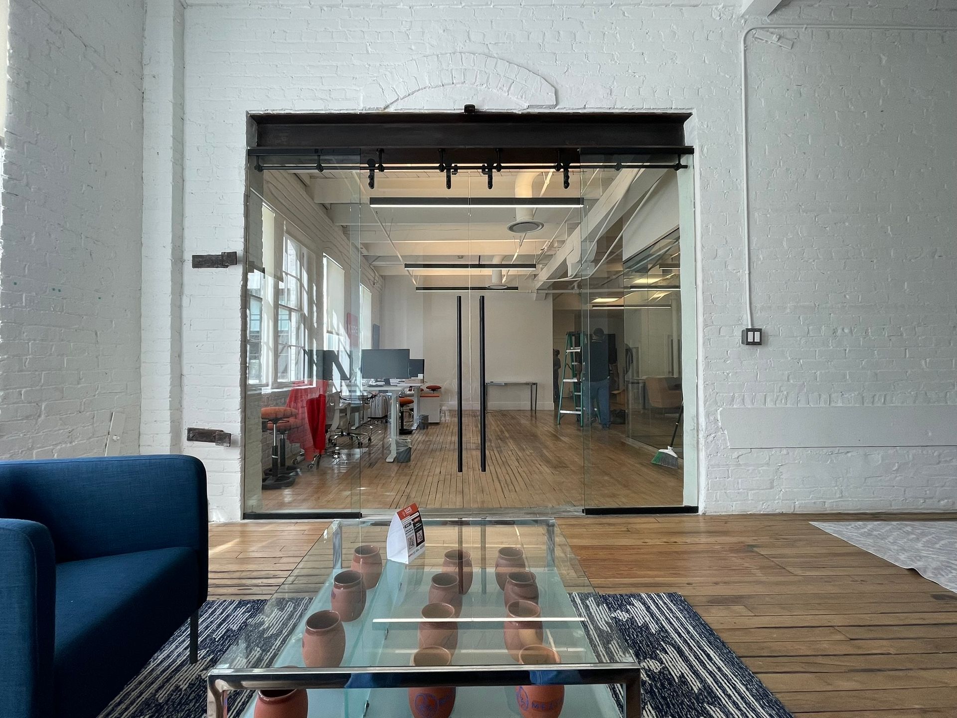A modern office interior featuring a blue couch, glass coffee table, and large glass double doors leading to a workspace.