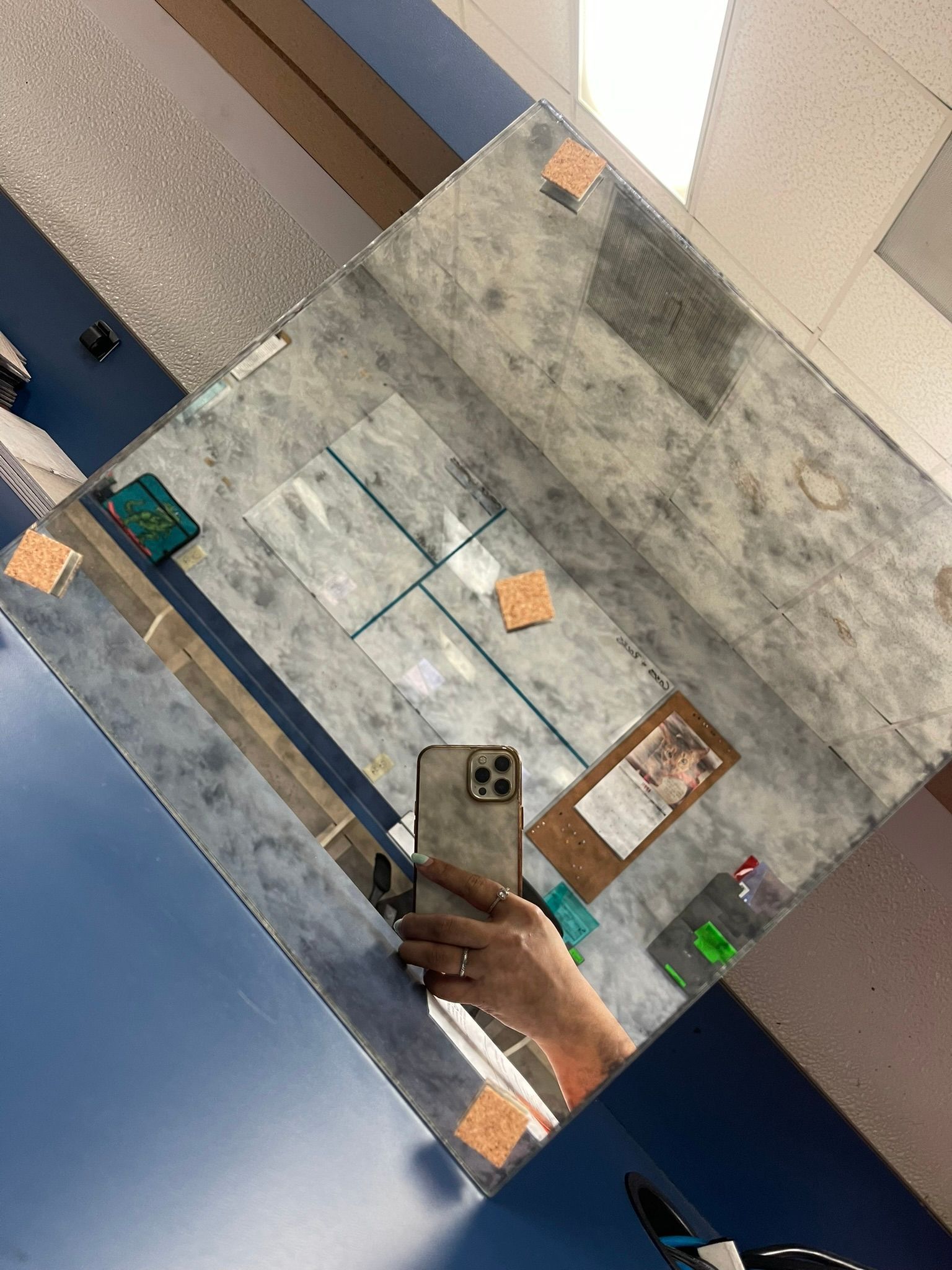 A person holds a phone to take a selfie in a square mirror with a mottled, gray, industrial-style surface.