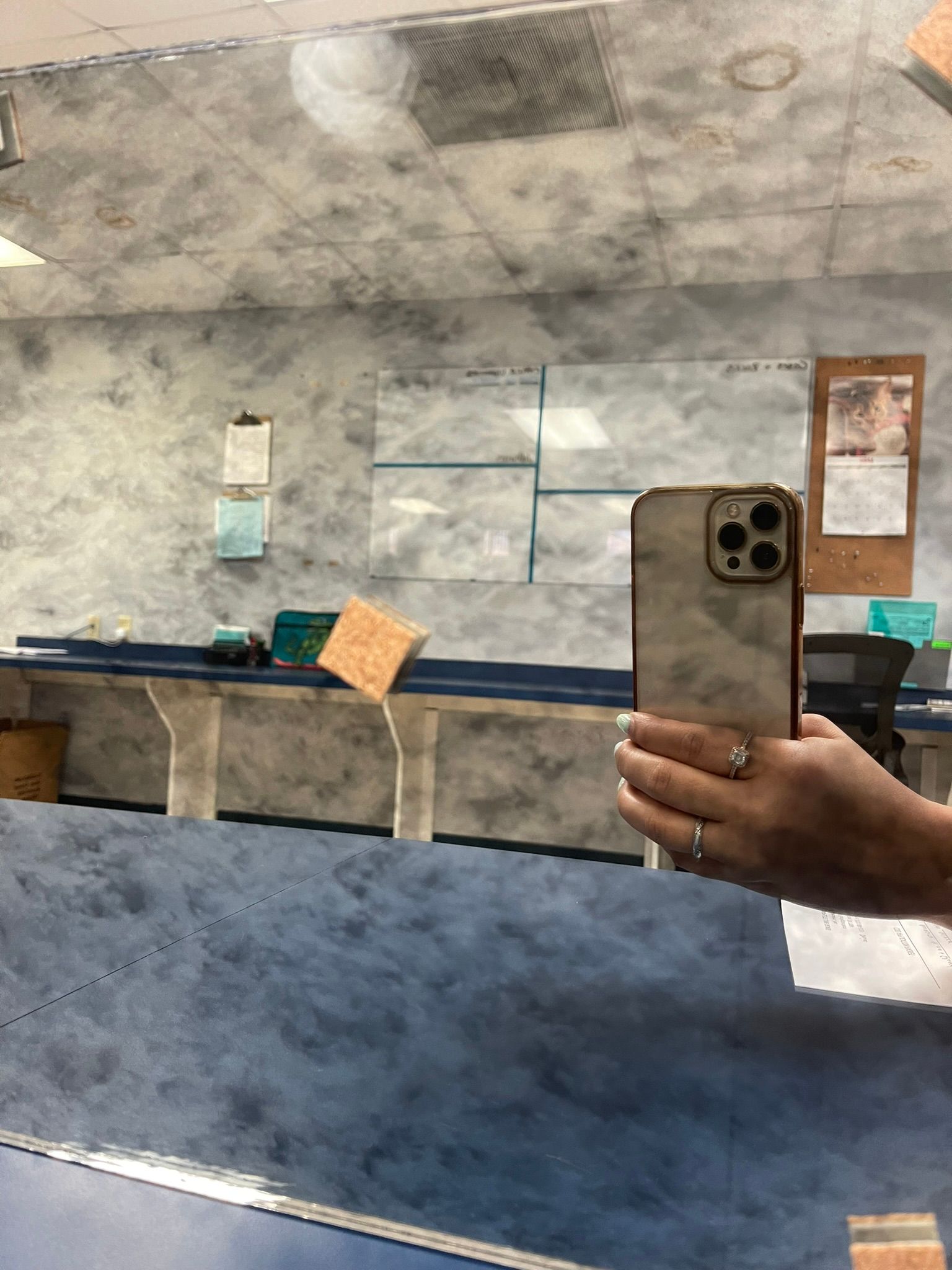 A mirror reflection showing a hand holding a phone, with a desk, office supplies, and a notice board in the background.