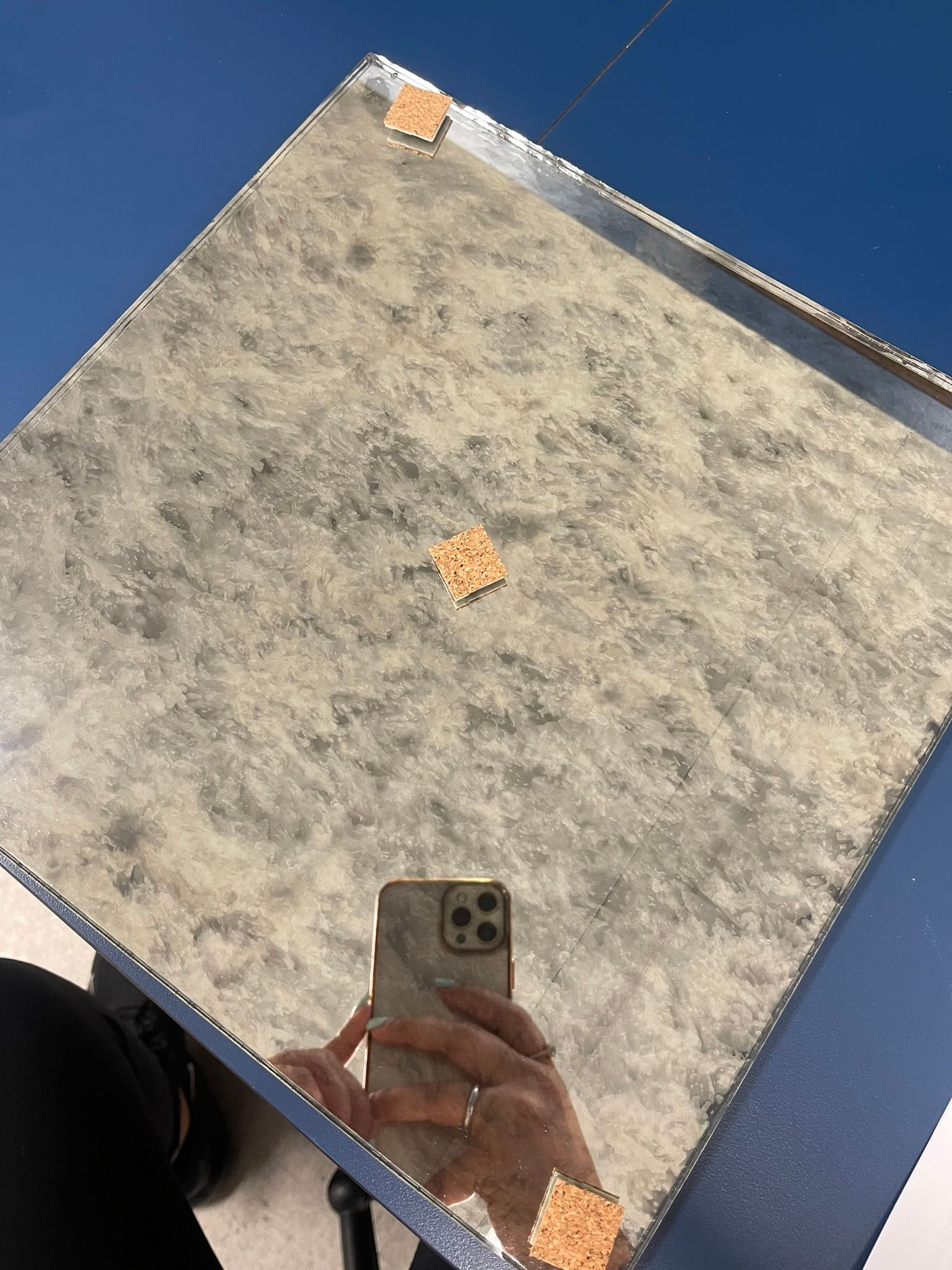 A person holds a phone to take a mirror selfie of a square, textured decorative tile with gold accents at the corners.