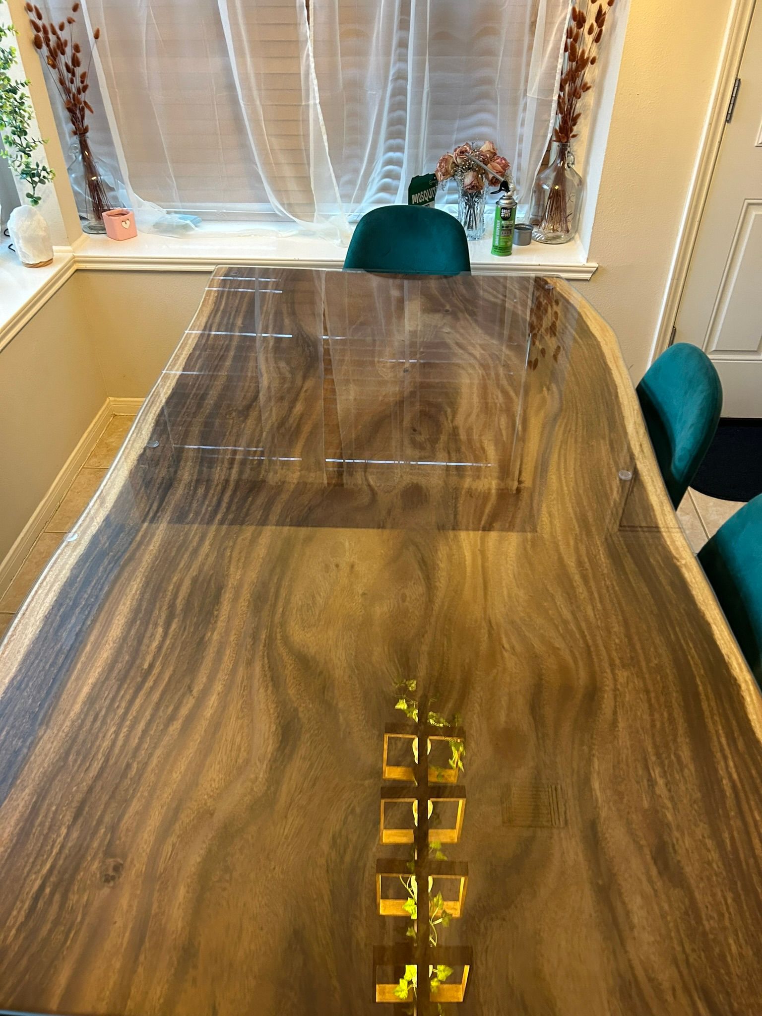 A polished wood dining table with a decorative gold-colored inlay sits in a sunlit room with three teal chairs.