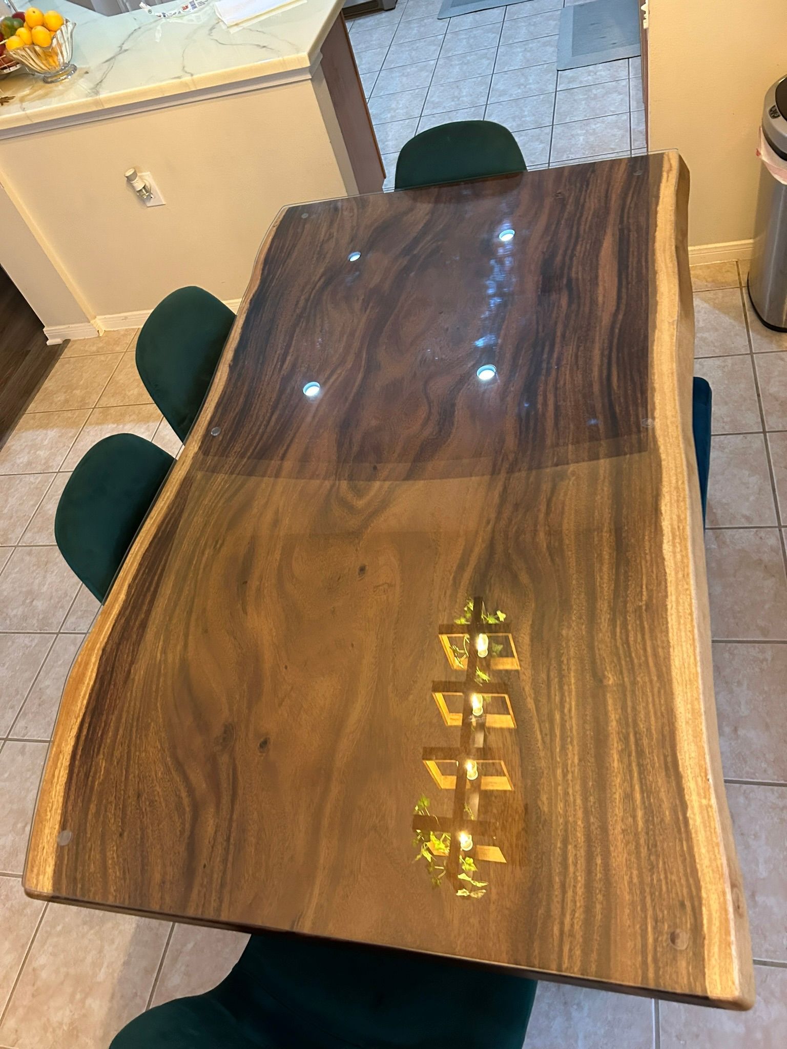 A live-edge wood dining table with a gold-colored, football-shaped resin inlay, viewed from above in a modern kitchen.