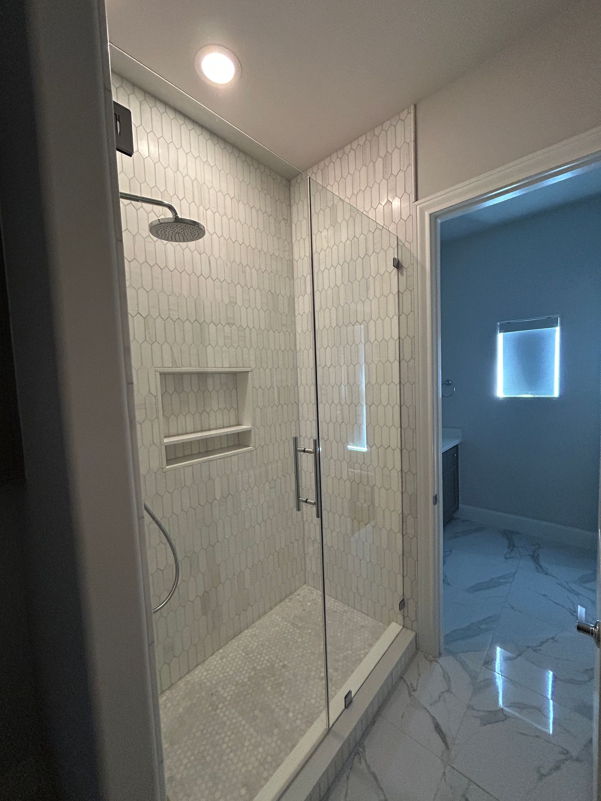 A walk-in shower with light-toned marble tile, a glass door, built-in wall niche, and a view into a bright bathroom.