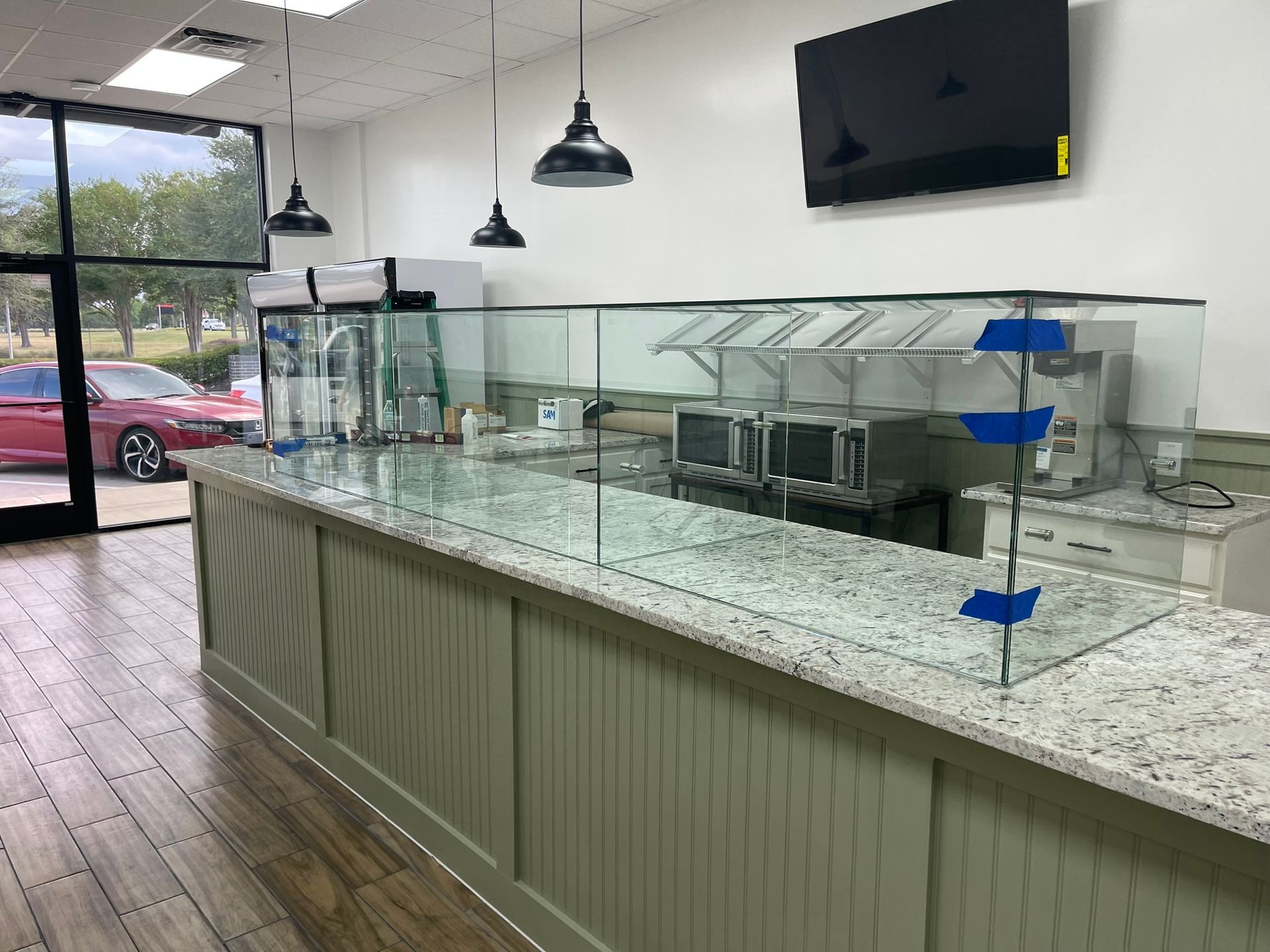 A service counter with a granite top and a glass sneeze guard, featuring green paneling and overhead industrial lighting.