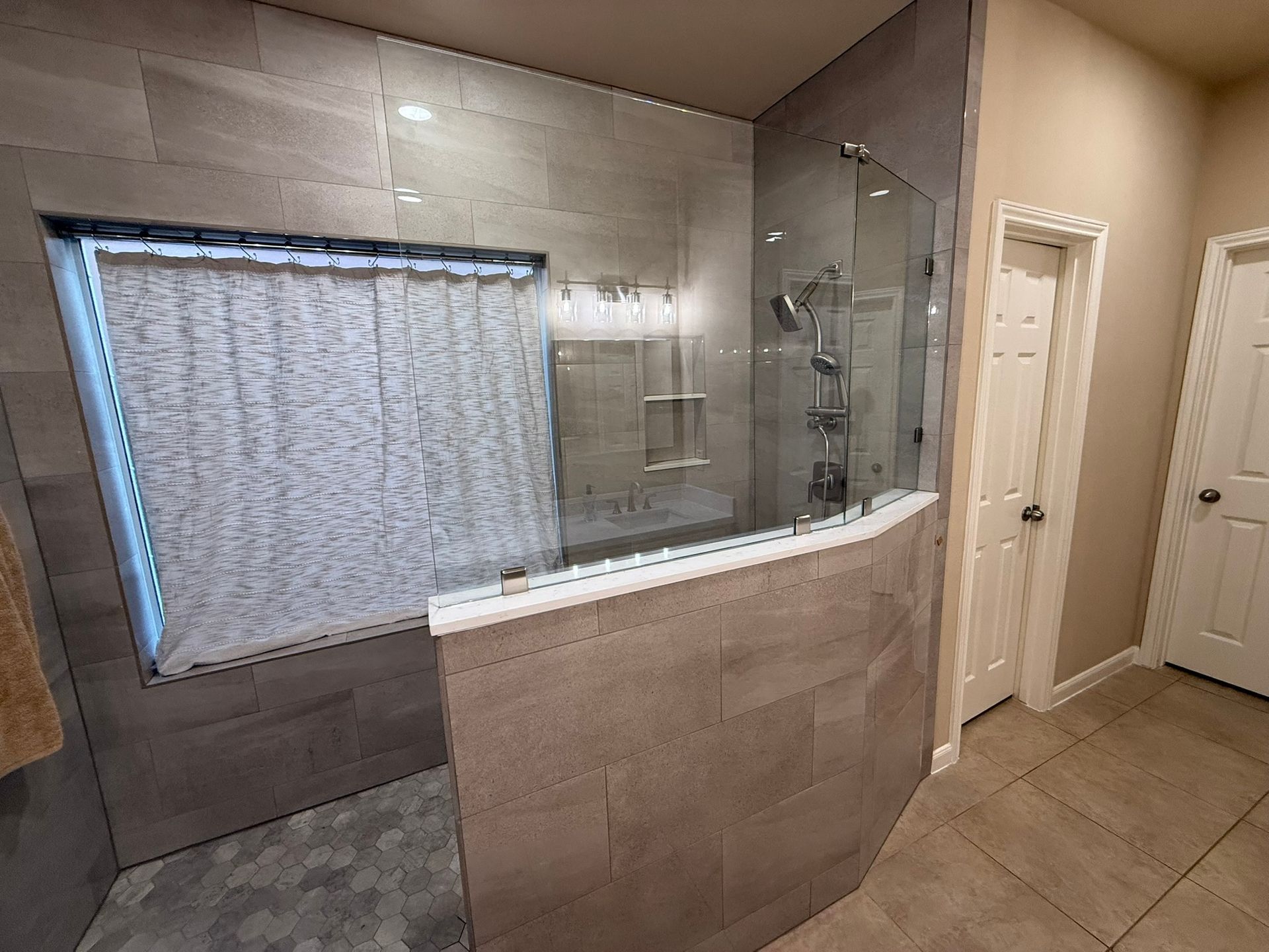 A modern bathroom shower area with grey stone-look tile, a glass partition, a window with blinds, and two white doors.
