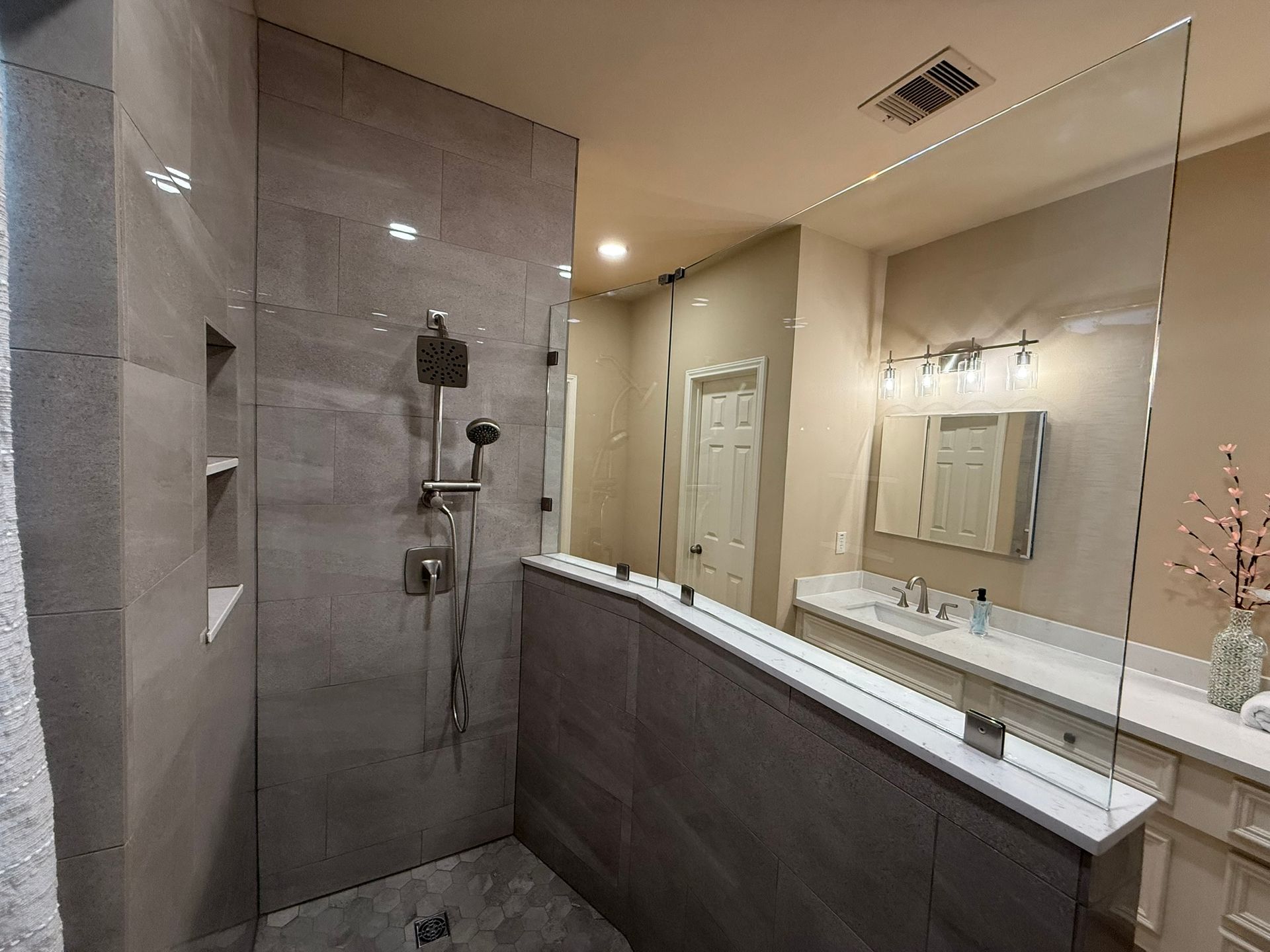A modern bathroom with a tiled walk-in shower, a half-wall partition, and a vanity mirror with overhead lighting.