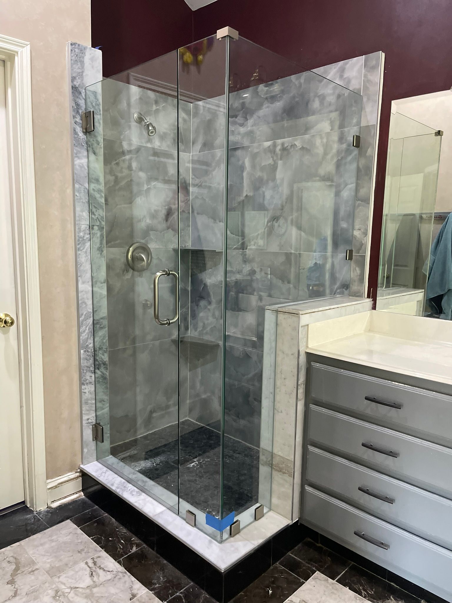 A glass-enclosed shower with grey marble tile, a chrome handle, and a black base, positioned next to a bathroom vanity.