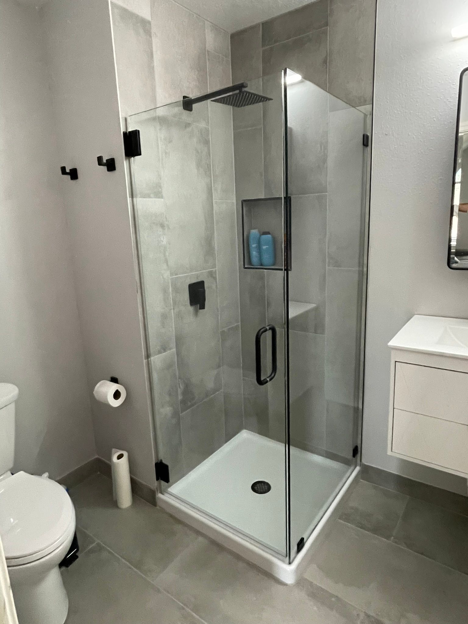 A modern bathroom with a glass-enclosed corner shower, grey tile walls, a white vanity, and a toilet.