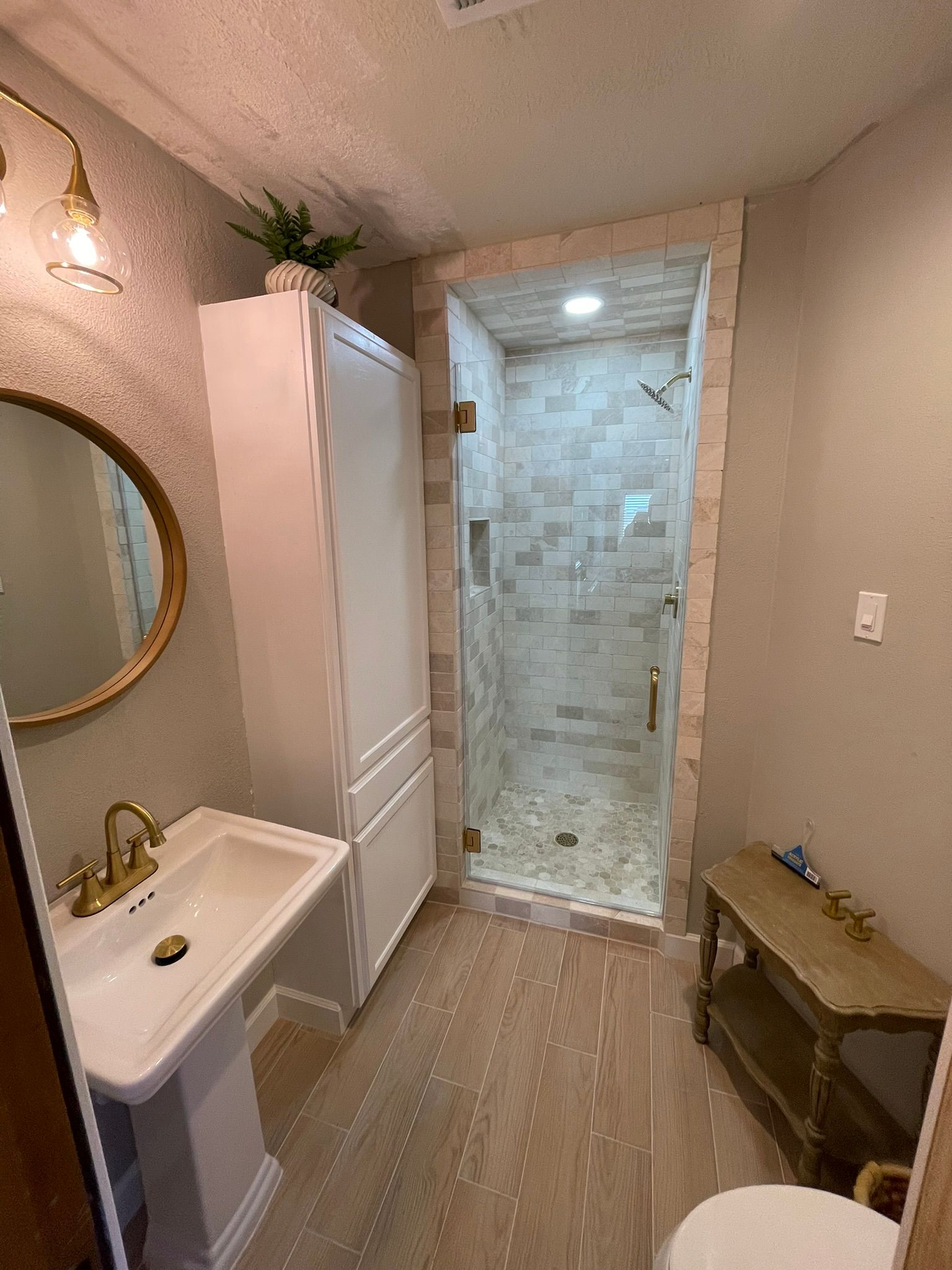 A bathroom featuring a pedestal sink, a tall white cabinet, a tiled walk-in shower, and a small wooden side table.