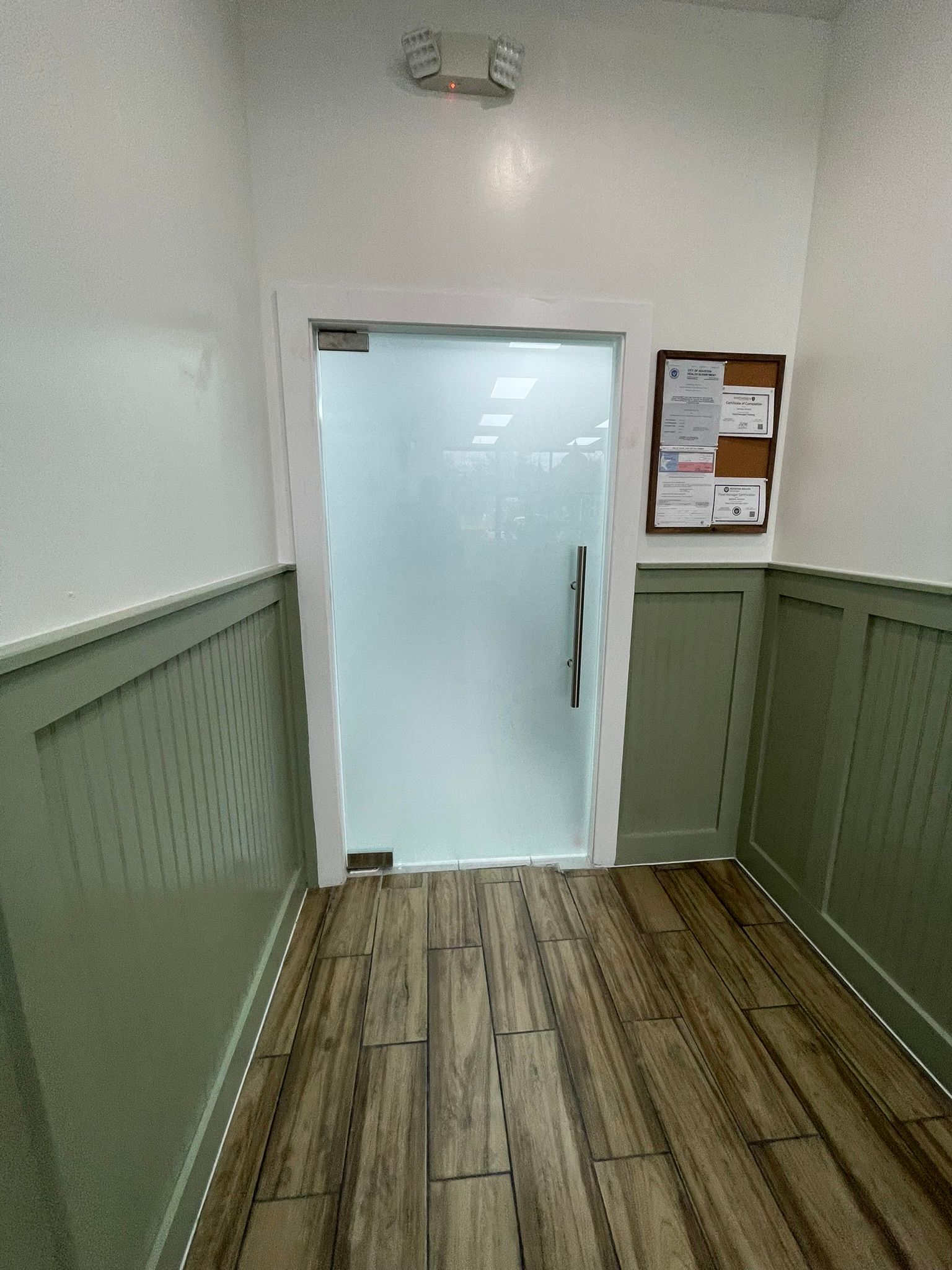 A narrow hallway with light green wainscoting and wood-look tile flooring leading to a frosted glass door.