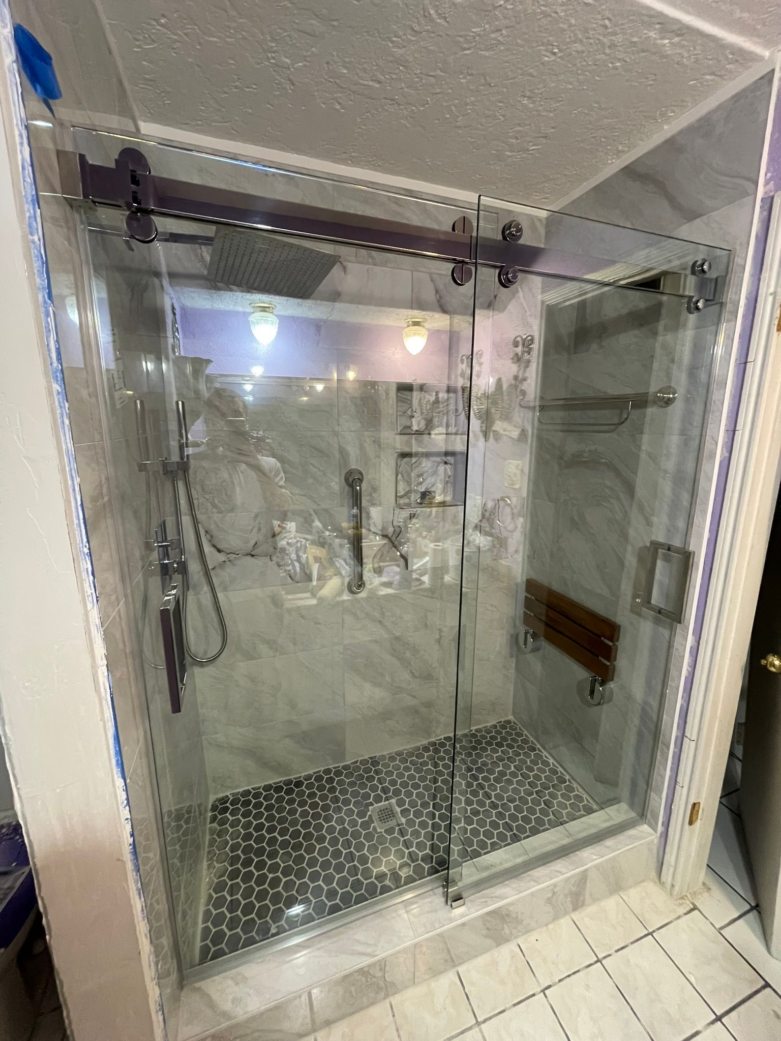 A glass walk-in shower with a sliding door, marble-patterned tile walls, a pebble-style floor, and a folding wood seat.