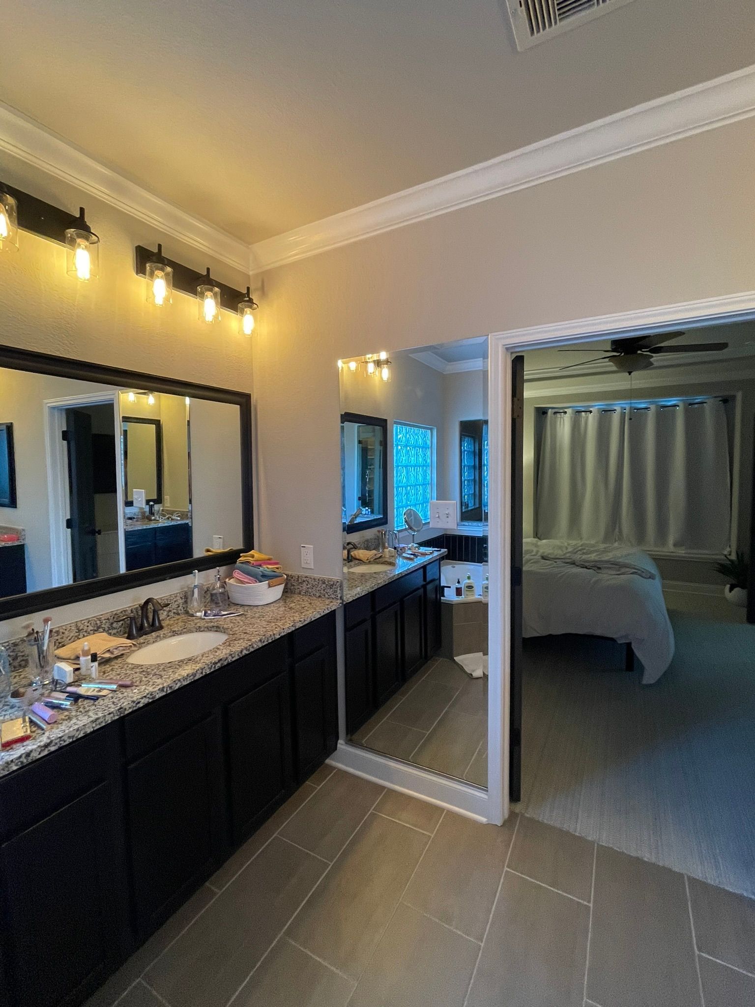 A modern bathroom vanity with dark cabinets and a large mirror, showing a reflection of an adjacent bedroom.