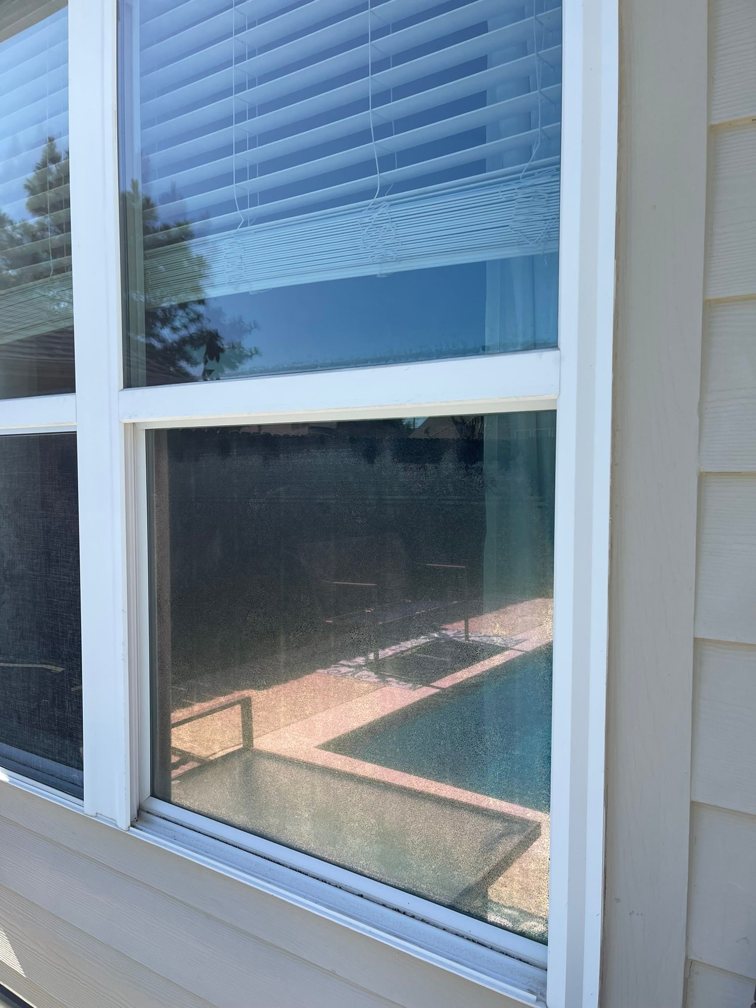 A bright white window frame installed in a tan siding wall, reflecting a backyard pool and patio on the glass.