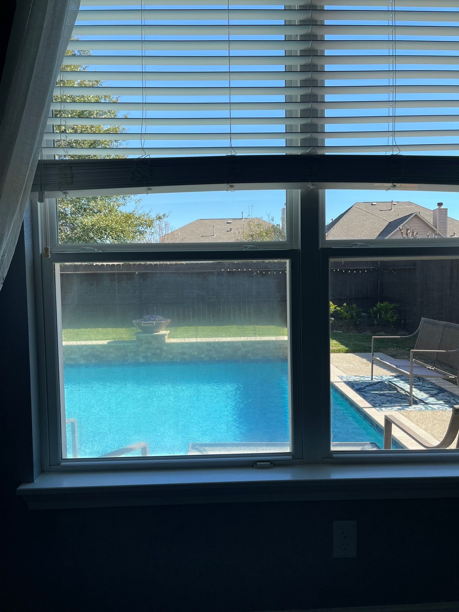 View of a backyard swimming pool and patio seen through a window with white blinds partially covering the top.