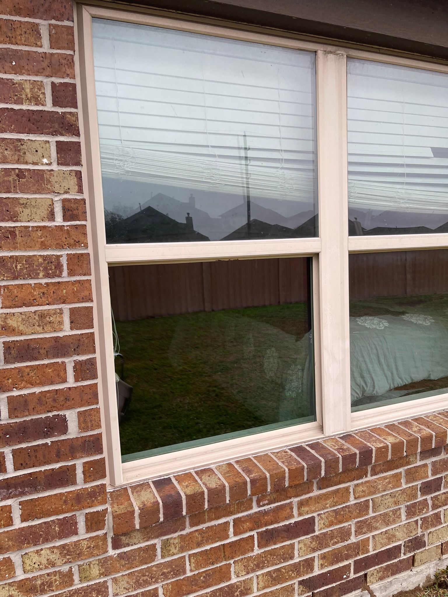 A close-up view of a beige-framed window set into a brown brick wall, showing a reflection of a fence and lawn.