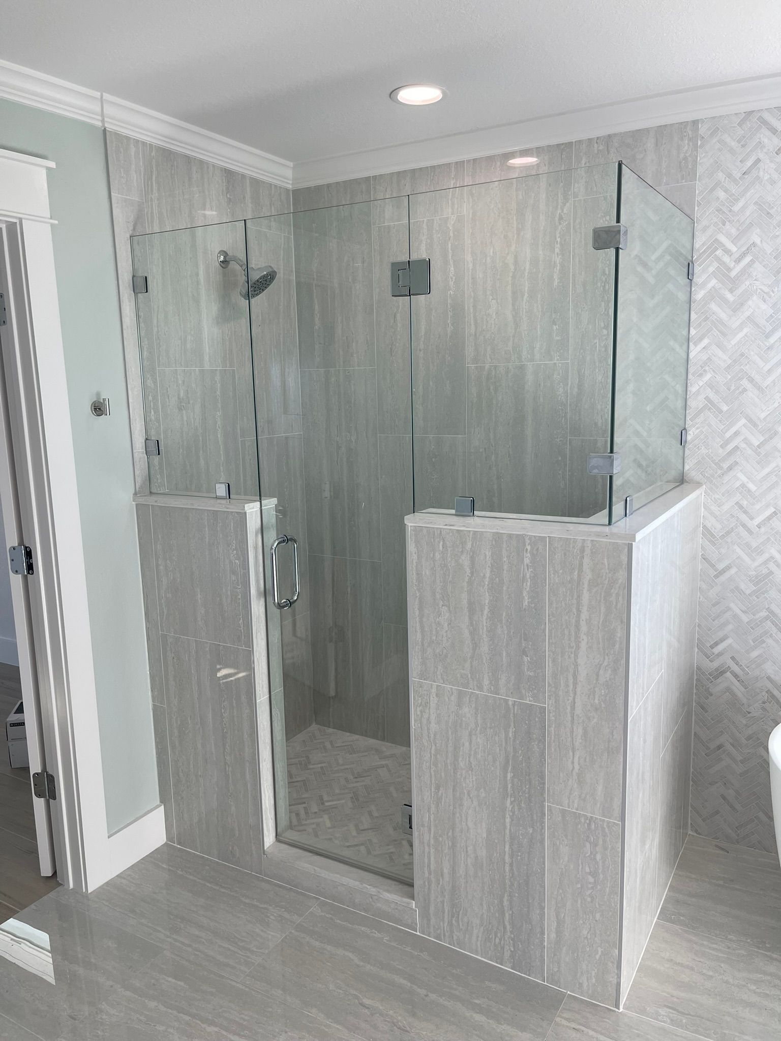 A modern walk-in shower with light gray tile walls, glass enclosure, and a half-wall entrance in a bathroom.