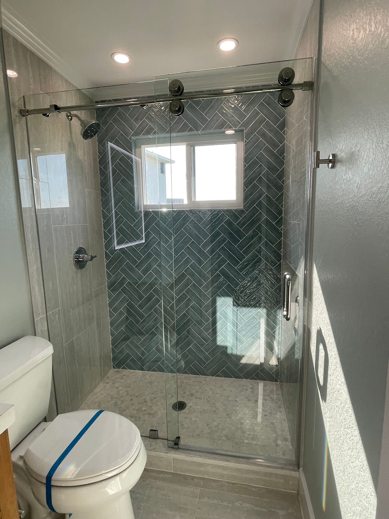 A glass-enclosed shower with dark herringbone tile walls and a light speckled floor, located in a bathroom.