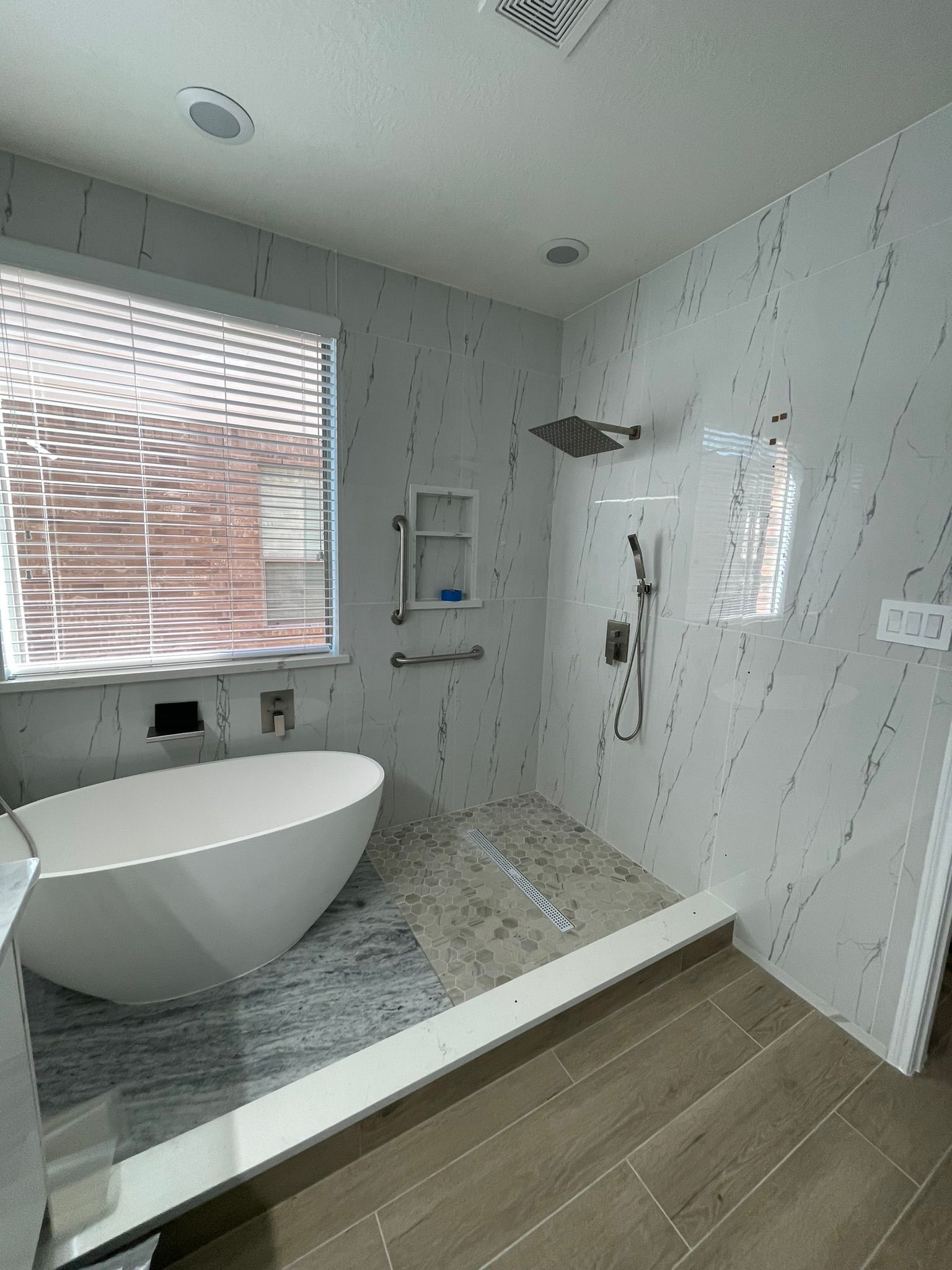 A modern bathroom featuring a white freestanding bathtub next to a large walk-in shower with marble tile and grab bars.