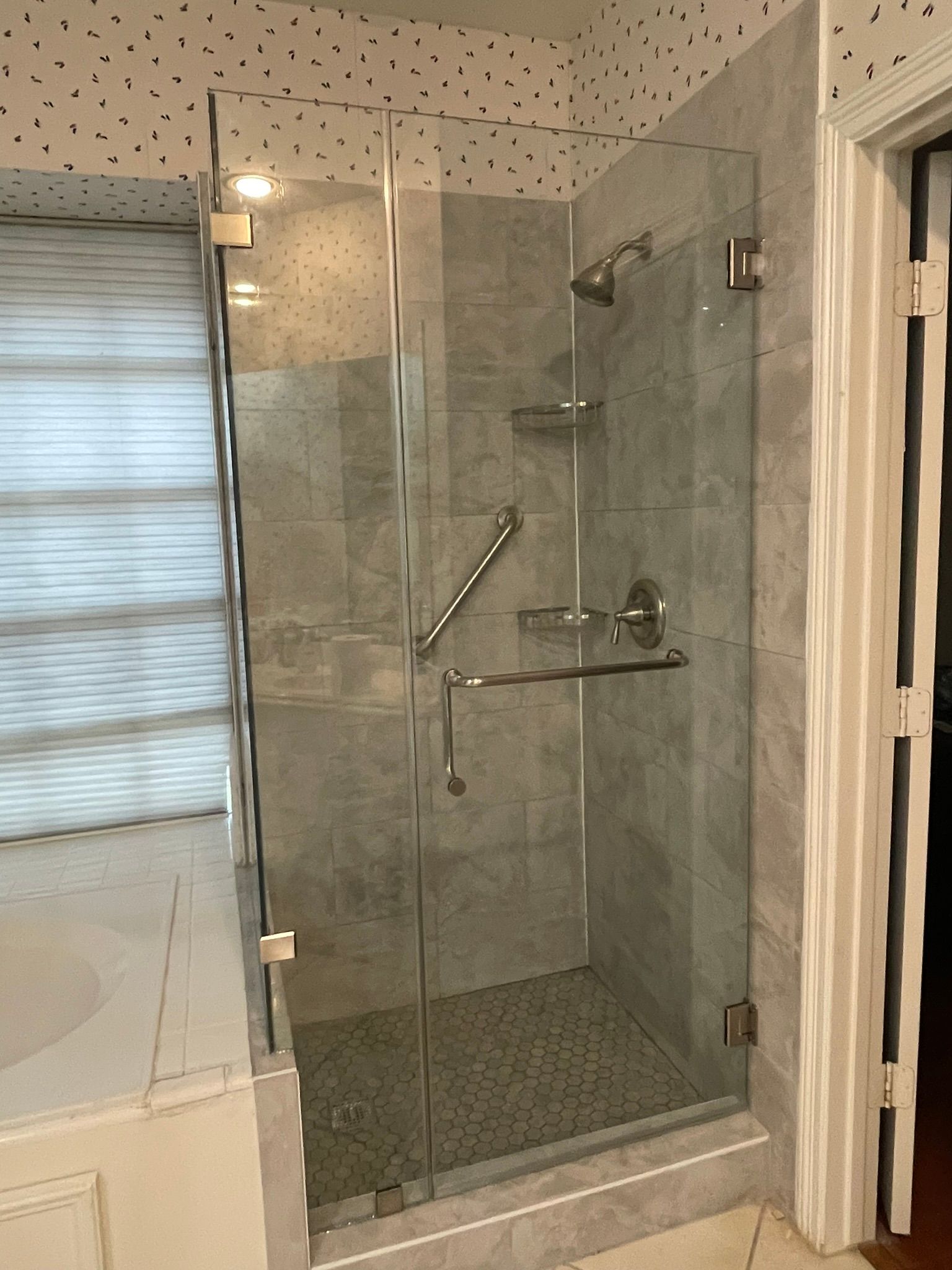A walk-in shower with a glass door, grey tiled walls, a built-in shelf, grab bars, and a patterned wallpaper border.