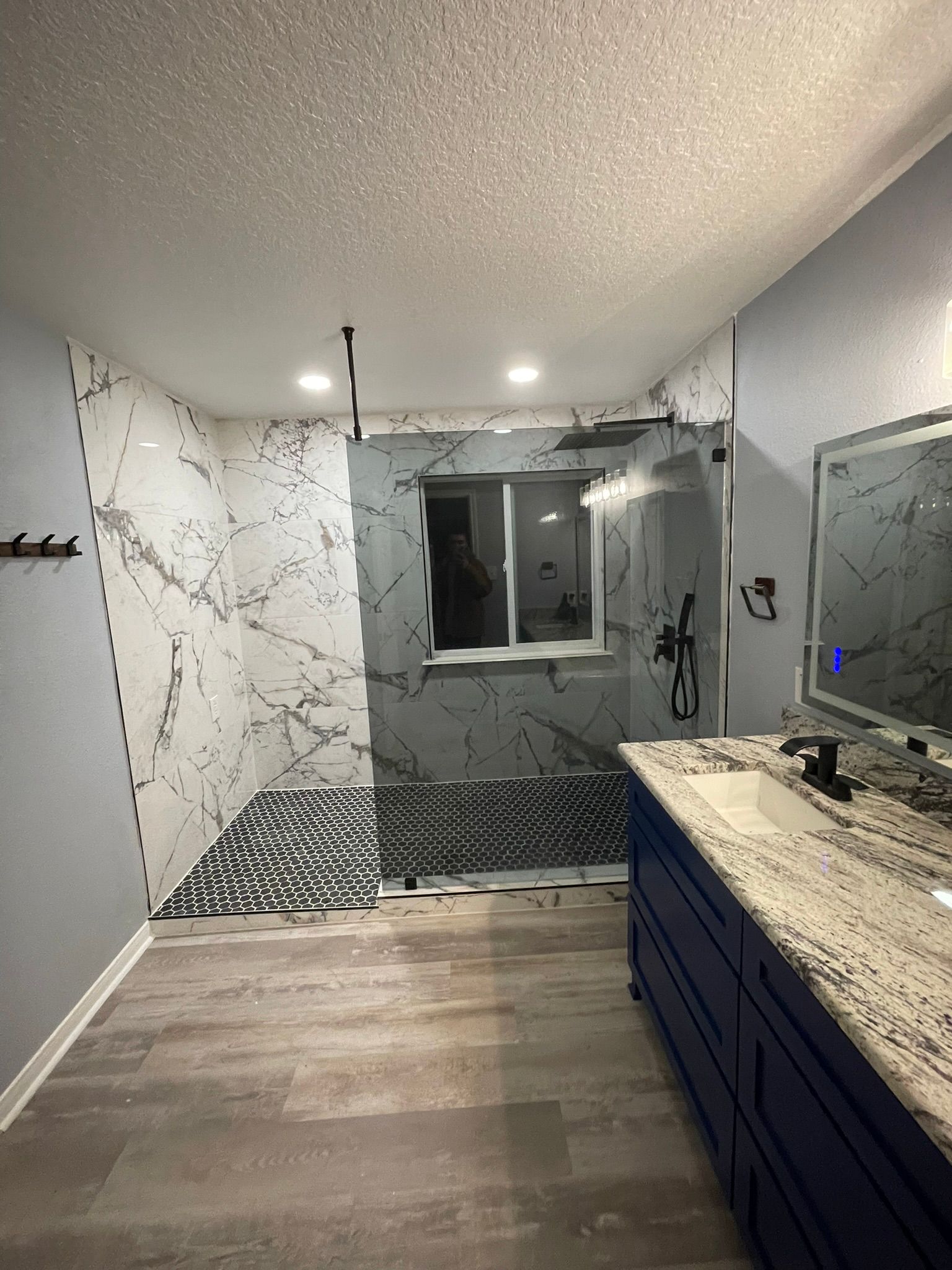 A bathroom under renovation featuring a white-and-gray marble tiled shower, dark blue vanity, and light gray wood flooring.