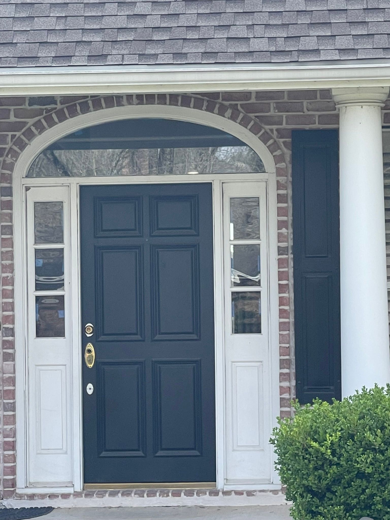 A dark blue six-panel front door with sidelights, a glass arched transom, and black shutters on a brick house exterior.