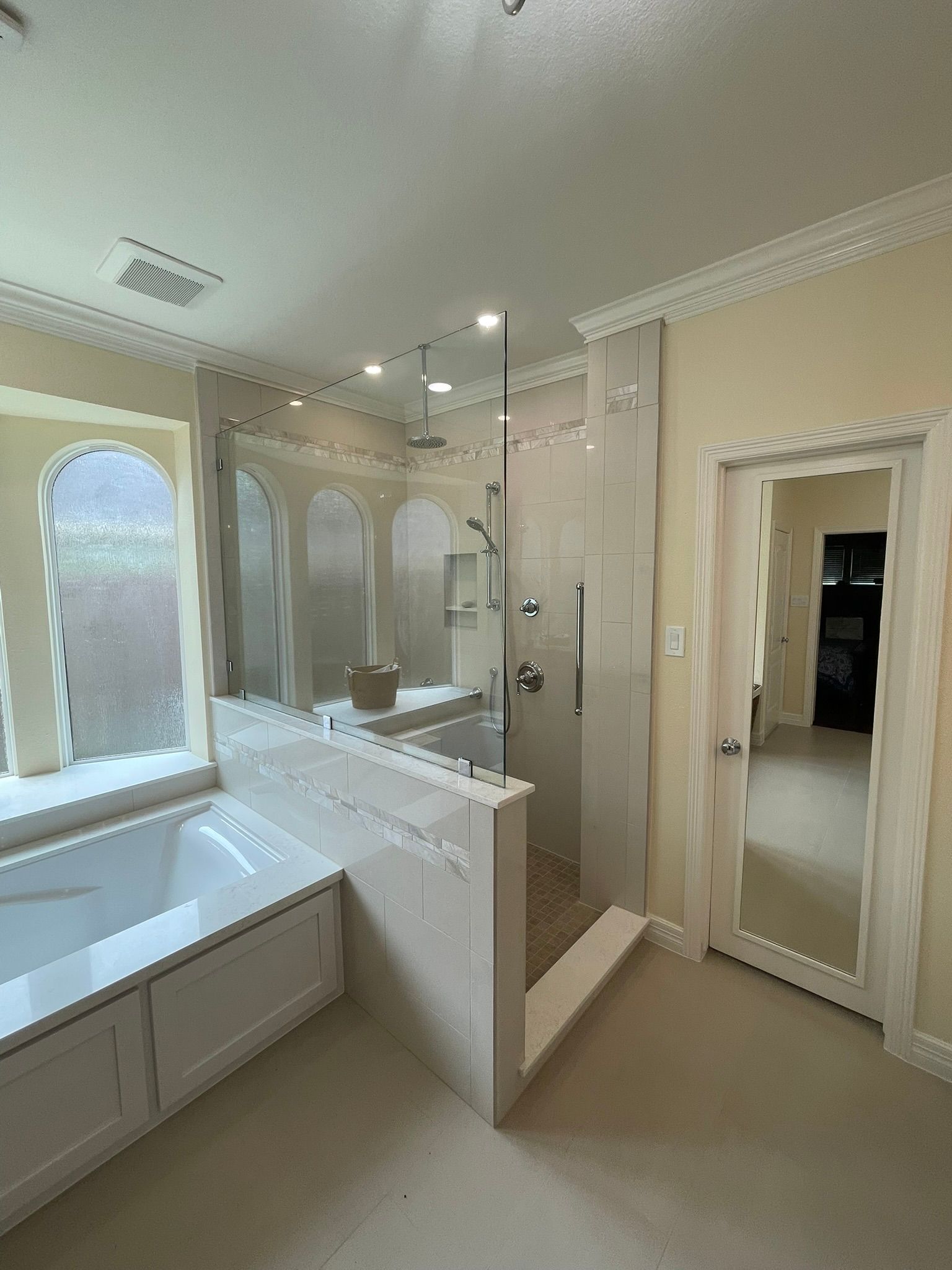 A bright, neutral-toned bathroom featuring a corner bathtub, a glass-enclosed walk-in shower, and an arched entryway.