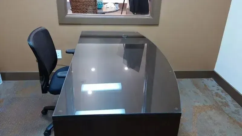 A modern, curved black desk with a glossy top sits in front of a dark brown wall, accompanied by a black office chair.