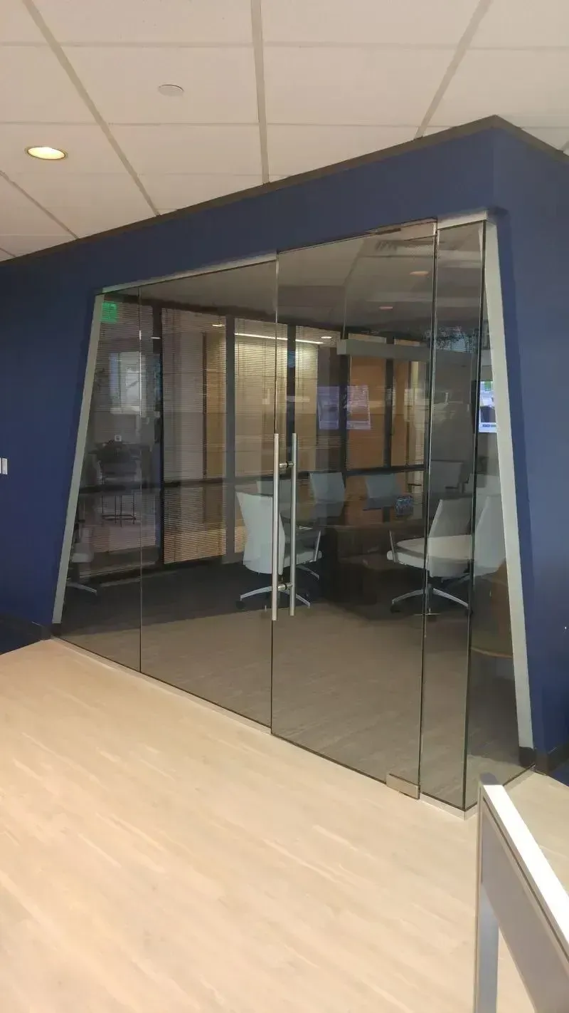 A glass-walled office conference room with a table and chairs set against dark blue walls in a modern office space.