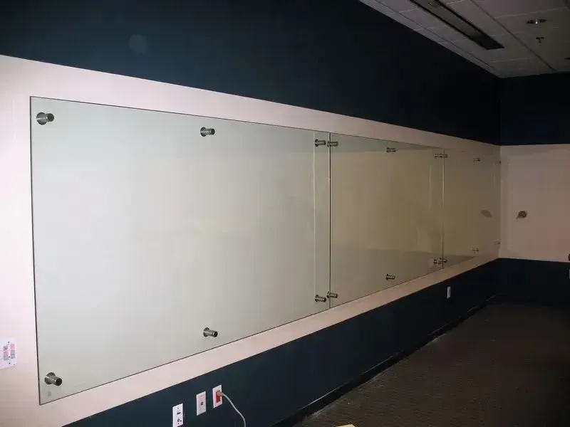 A wall-mounted glass whiteboard featuring metal standoffs on a wall with dark and light-colored paint sections.