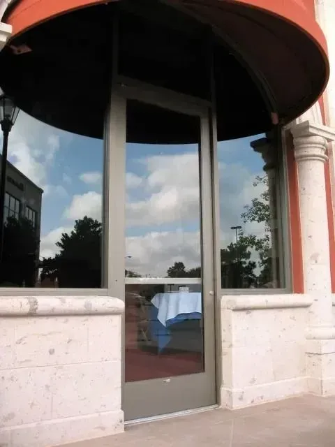 A glass storefront door framed by a stone wall under a red awning, reflecting the cloudy sky and an indoor table setting.