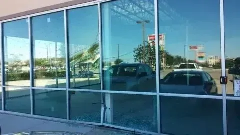 A modern building storefront with large glass windows reflecting a parking lot, cars, and a clear blue sky.