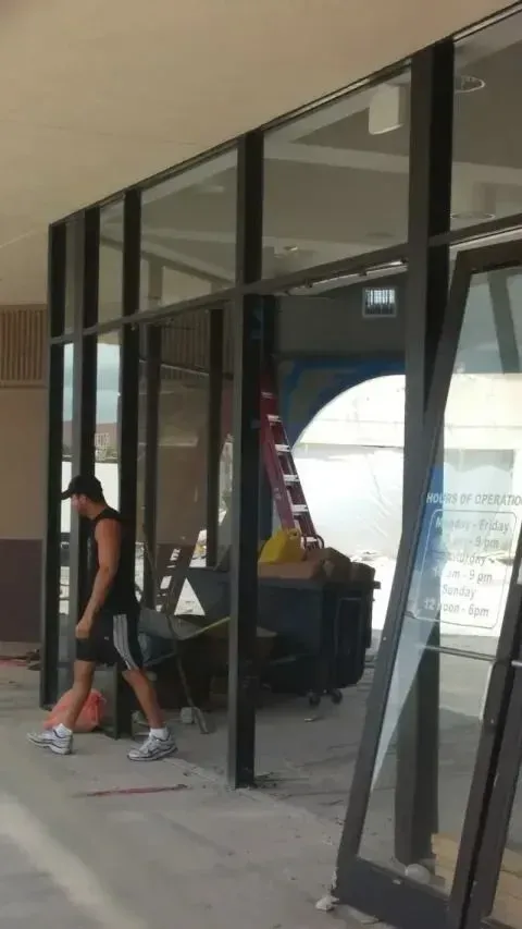 A person in a black tank top and shorts walks past the large glass windows of a storefront under construction.