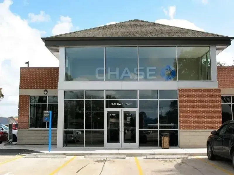 A Chase bank branch with a brick exterior, large glass windows, and a parking lot in front under a blue sky.