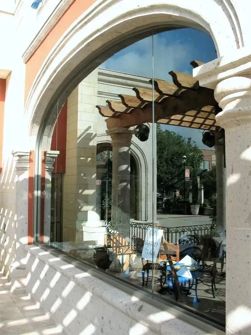A window reflects a stone pergola and outdoor seating area on a sunny day with distinct striped shadows on the ground.