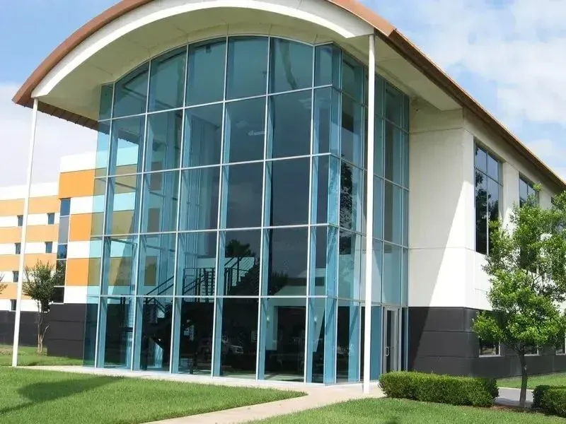 A modern two-story building features a prominent glass curtain wall facade under a curved roof, surrounded by lawn.