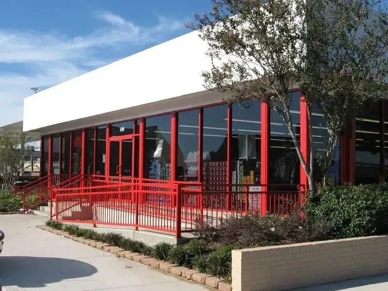 A commercial building with a white facade, red window frames, and a red metal wheelchair ramp leading to the entrance.