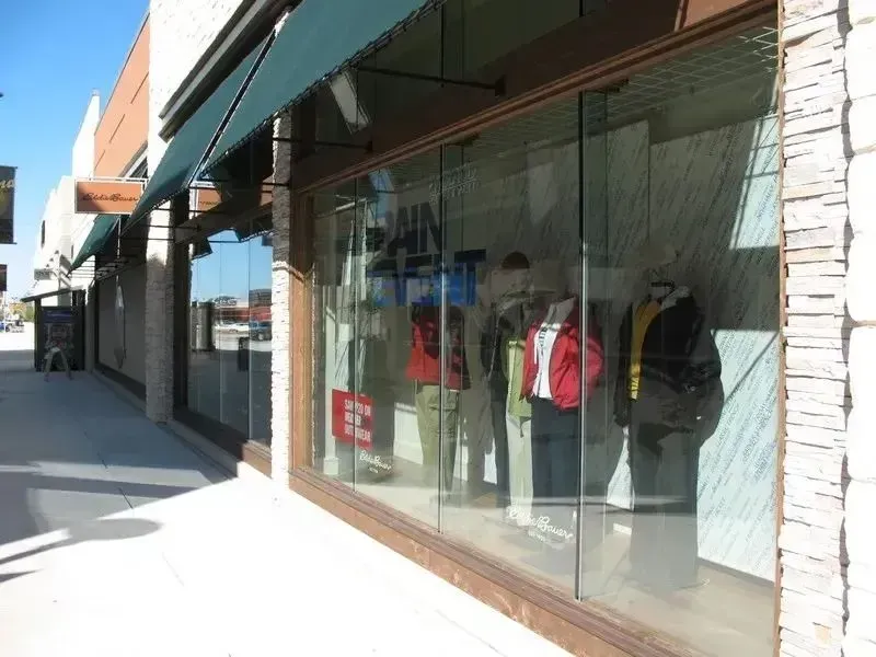 A storefront window displays several mannequins dressed in casual clothing, situated under a green awning on a sunny day.