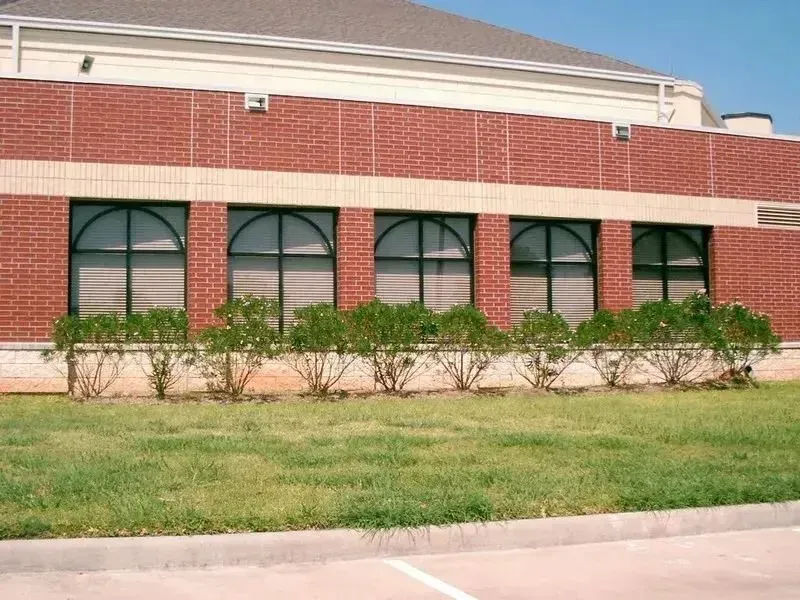 A brick building wall with five arched windows above a row of bushes and a grassy lawn.
